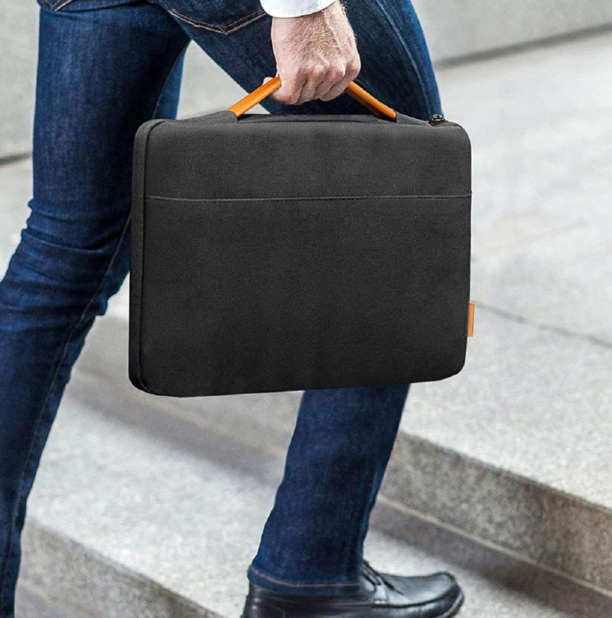 Person in Jeans, die eine schwarze Laptoptasche mit orangefarbenem Griff trägt.