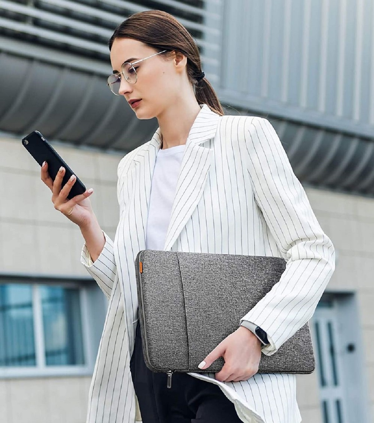 Frau im Blazer hält eine graue Laptoptasche und ein Telefon, steht vor einem modernen Gebäude.