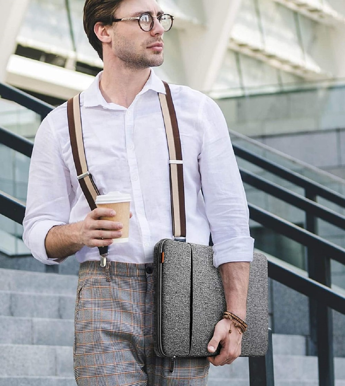 Ein Mann in einem weißen Hemd und Hosenträgern hält einen Kaffeebecher und eine graue Laptoptasche.