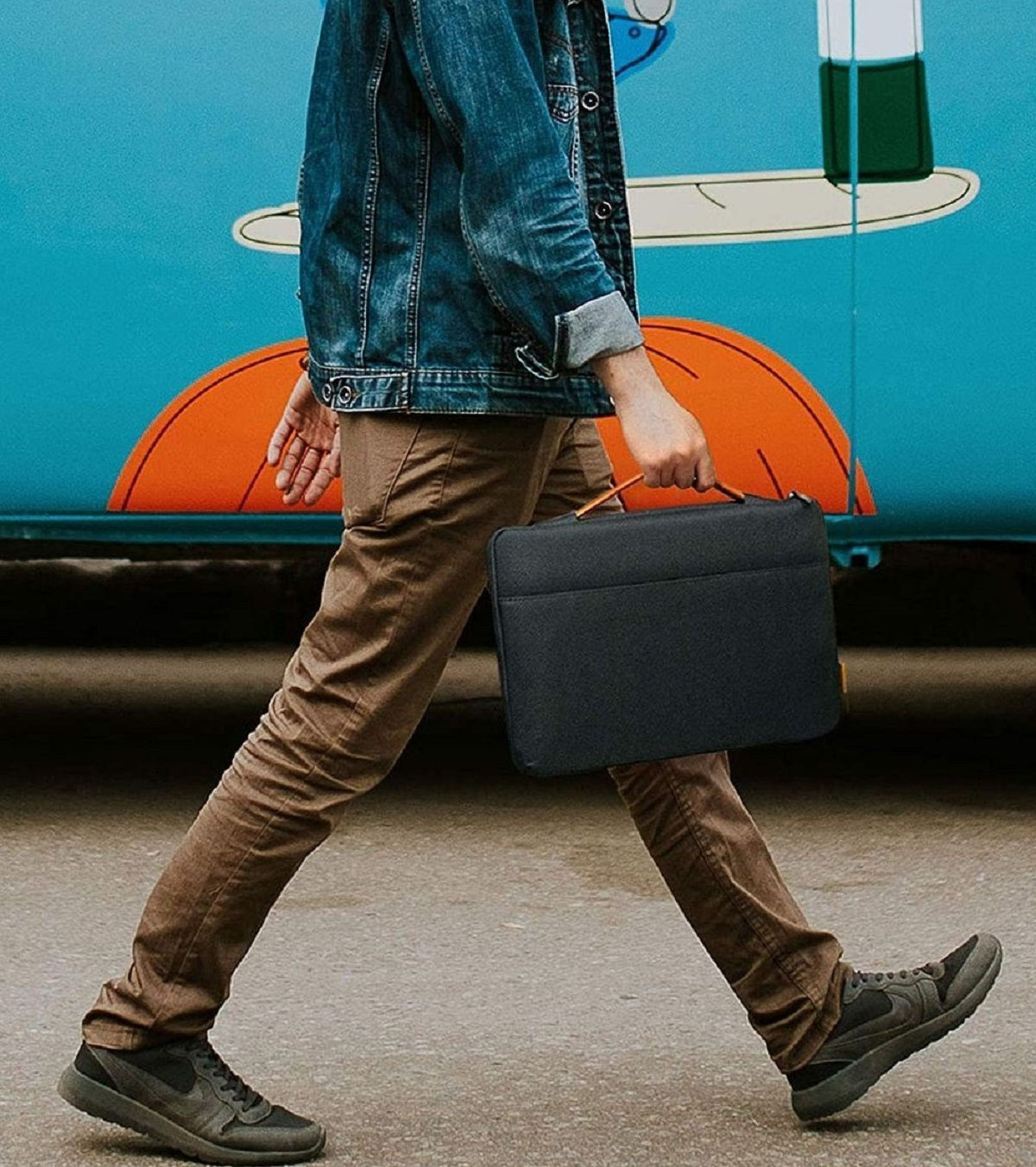 Mann in Jeansjacke und brauner Hose geht mit schwarzer Laptoptasche vor blauem Bus.