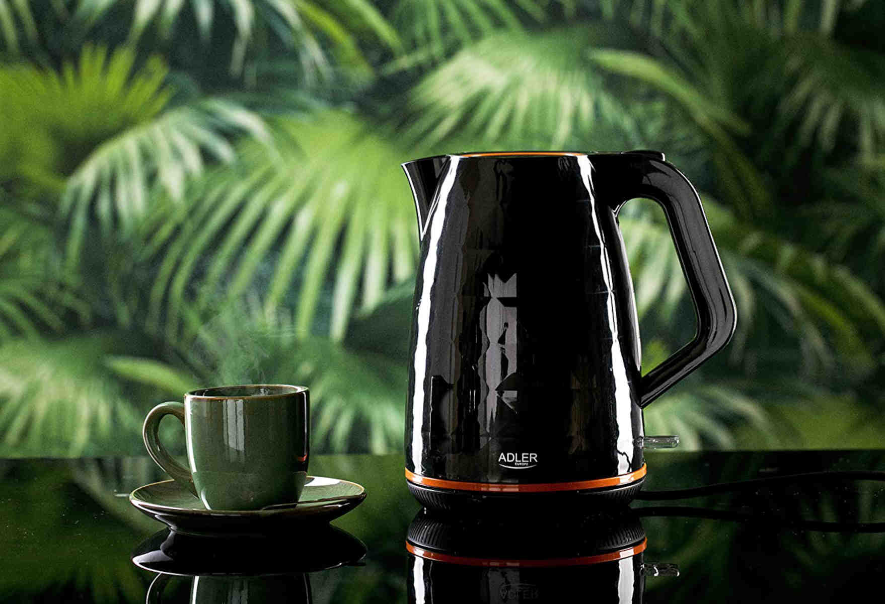 Bouilloire noire avec des accents oranges, une tasse et une soucoupe sur une surface réfléchissante.