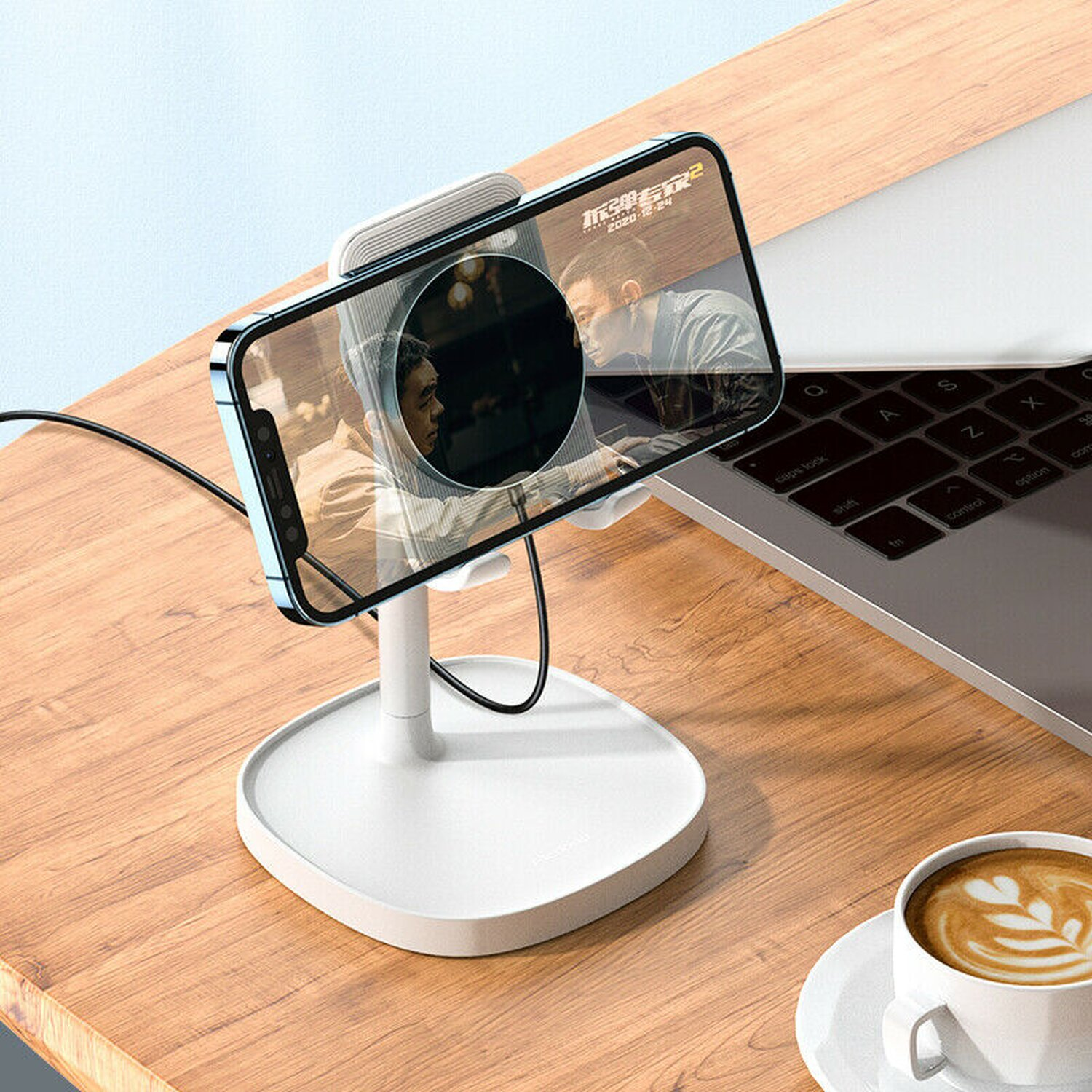 Ein Smartphone, das einen Film zeigt, auf einem weißen Ständer mit angeschlossenem schwarzem Kabel. Eine Tasse Kaffee im Hintergrund.