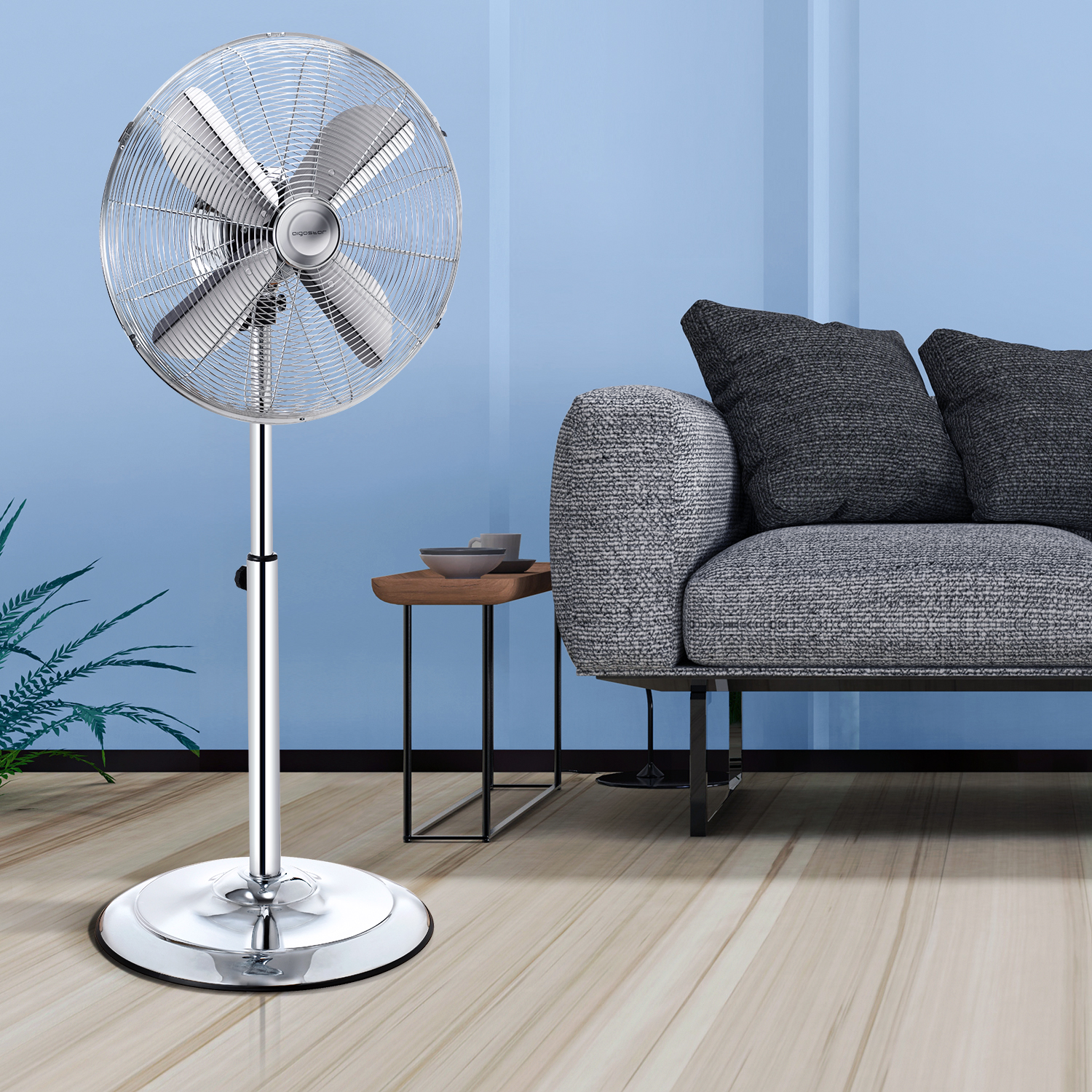 Un ventilatore a colonna cromato si trova in una stanza con pareti azzurro chiaro e un divano grigio.