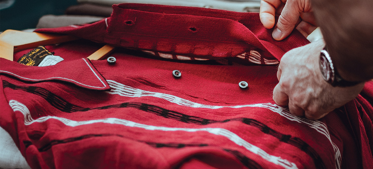 Le mani di una persona sistemano una camicia rossa a righe bianche. Appendiabiti in legno visibile.