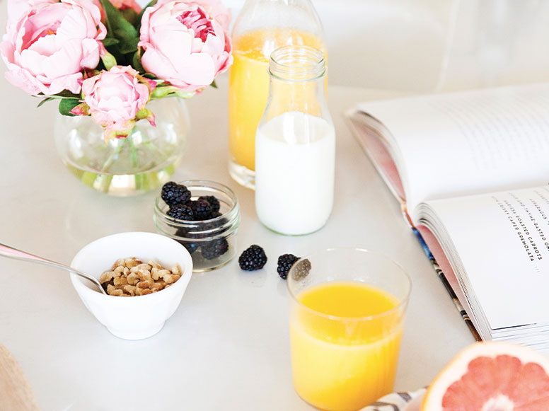 Stół śniadaniowy z kwiatami, sokiem, mlekiem, płatkami, jeżynami i książką. Jasne, naturalne oświetlenie.