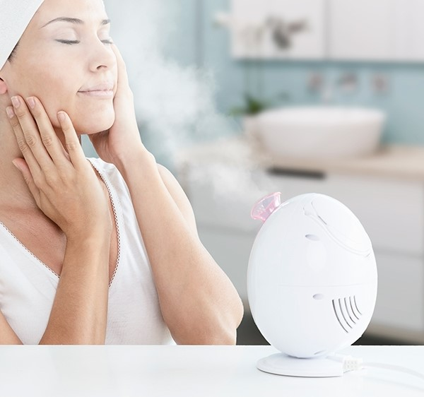 Donna che usa un vaporizzatore facciale in bagno, con gli occhi chiusi. Dispositivo bianco visibile.