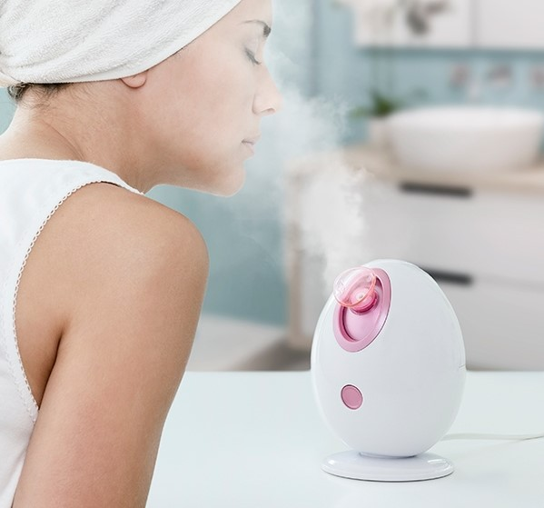 Una donna che fa il vapore al viso con un vaporizzatore facciale. Ha un asciugamano sulla testa.