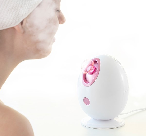 Donna che usa un vaporizzatore facciale, bianco con accenti rosa, vapore visibile.