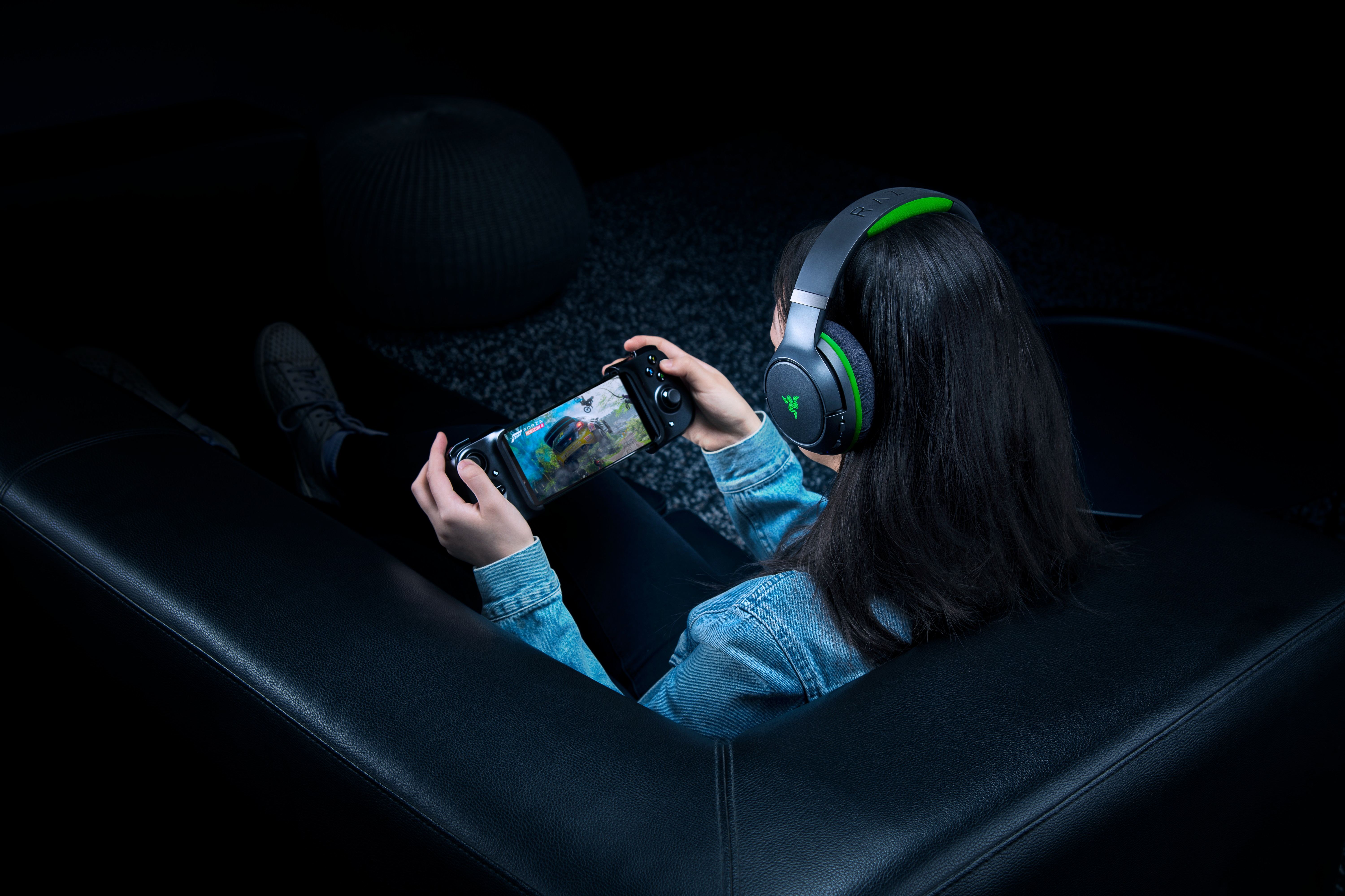 Eine Person auf einer schwarzen Couch, die Kopfhörer trägt und einen Gamecontroller mit Telefon hält.