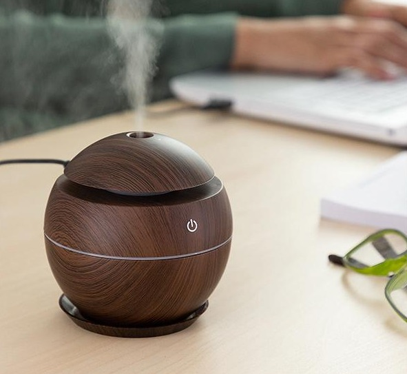 Un difusor de aceites esenciales con aspecto de madera sobre un escritorio, con vapor, un portátil y una mano al fondo.