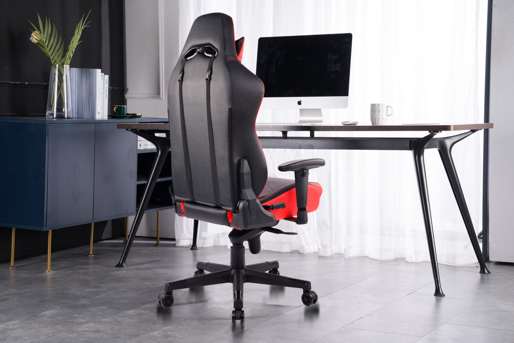 Ein Gaming-Stuhl steht in einem Raum mit Schreibtisch, Computer und Pflanzen. Der Stuhl ist schwarz und rot.
