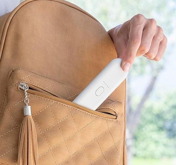 Eine Hand legt ein weißes Gerät in die Tasche eines beige-farbenen Rucksacks mit Quaste.