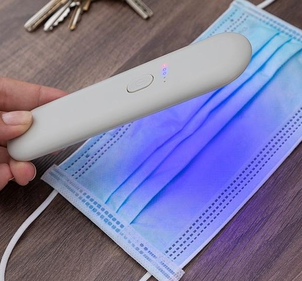 Eine Hand hält ein weißes UV-Licht über einer blauen OP-Maske auf einem Holztisch.