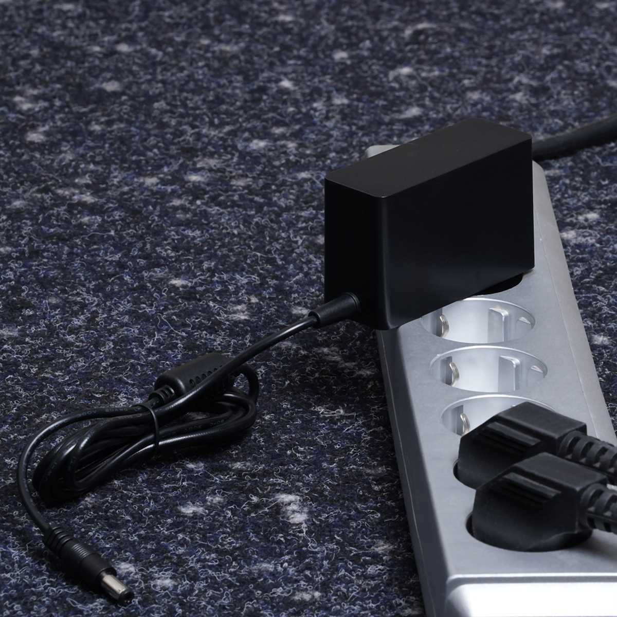 Ein schwarzes Netzteil ist an einem Steckdosenleiste angeschlossen. Der Hintergrund ist ein grauer Teppich.