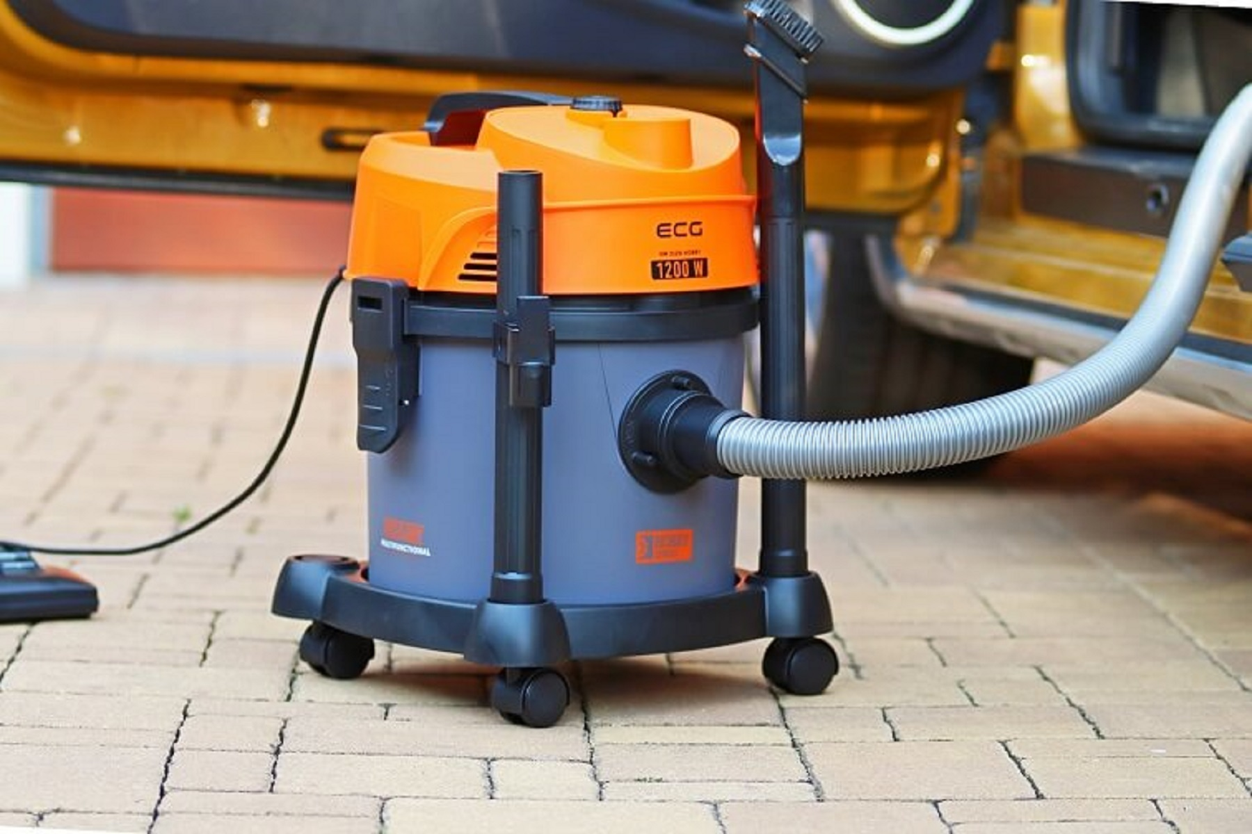 Un aspirapolvere con corpo grigio e parte superiore arancione su una base con ruote. Un tubo si estende verso un'auto.