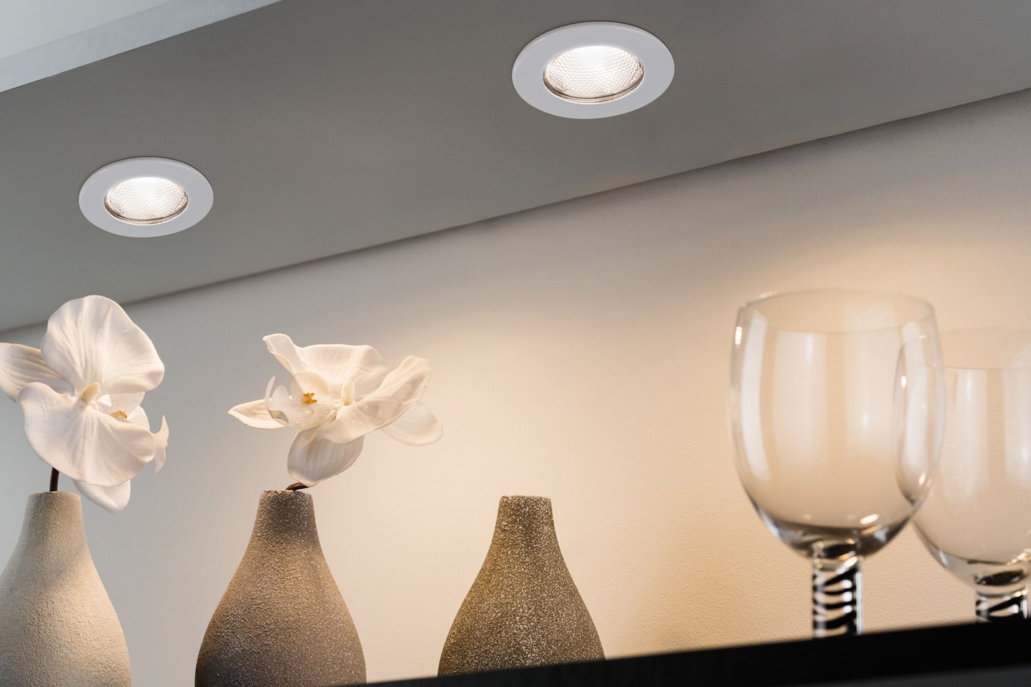 Zwei Einbauleuchten in einer Decke, die weiße Vasen mit Blumen und Weingläser beleuchten. Licht und Schatten.