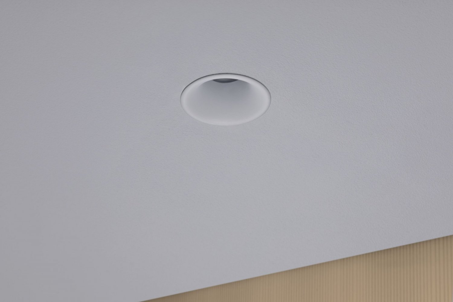 Ein Einbaustrahler, kreisförmig, an einer weißen Decke. Unten befindet sich eine beige Wand.