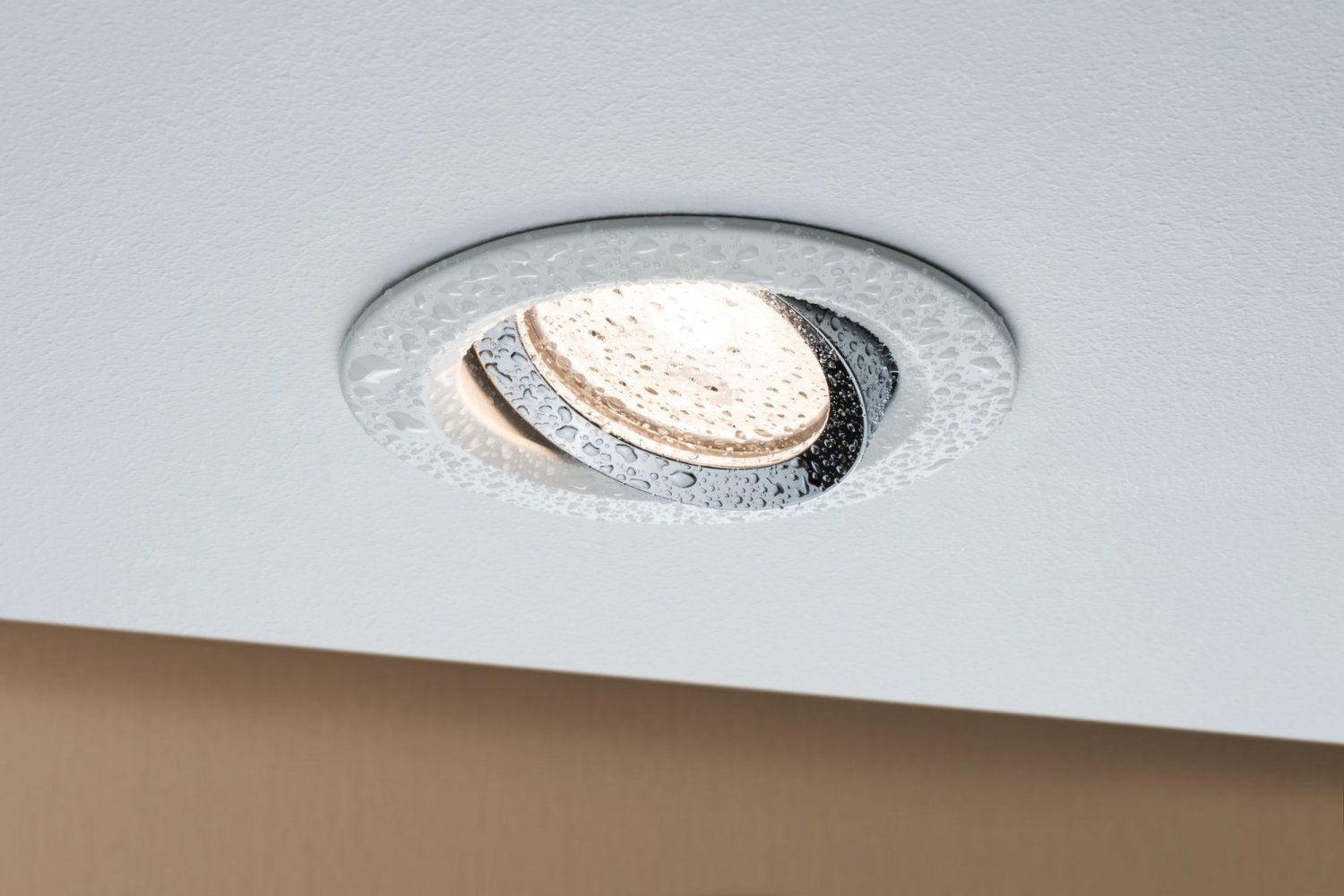 Deckenleuchte mit Chromring, Wassertropfen und beleuchteter Mitte. Braune Wand im Hintergrund.