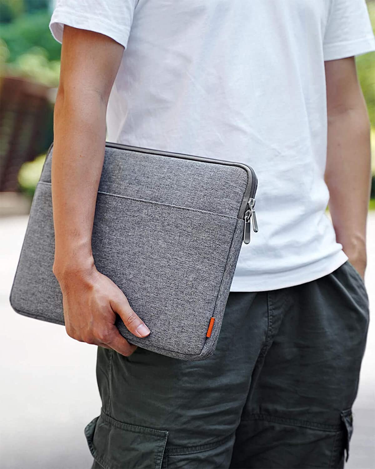 Hombre sosteniendo una funda gris para portátil, camisa blanca y pantalones cargo. Fondo verde.