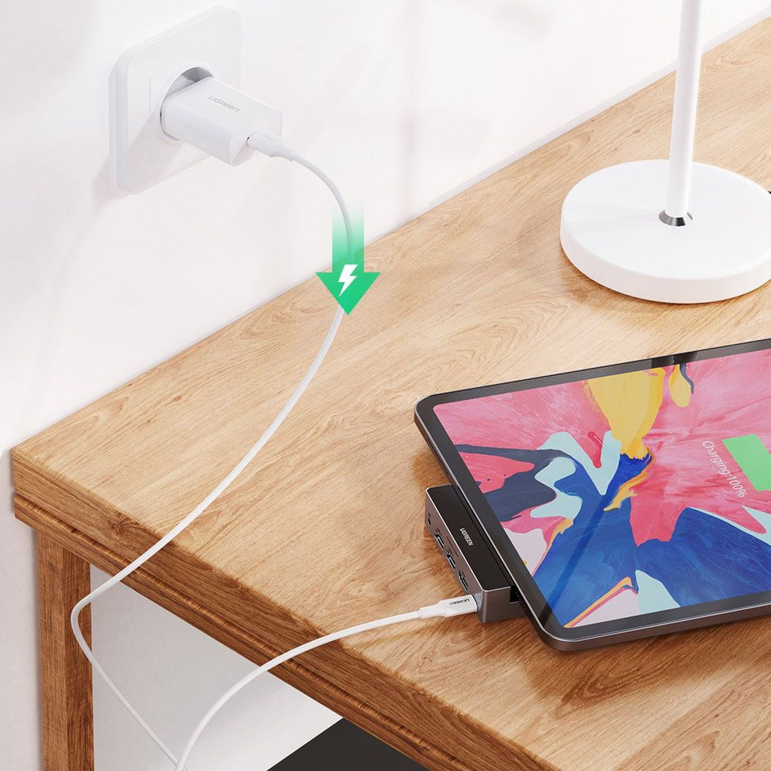 A phone charging on a wooden table with a charger, a tablet, and a desk lamp.