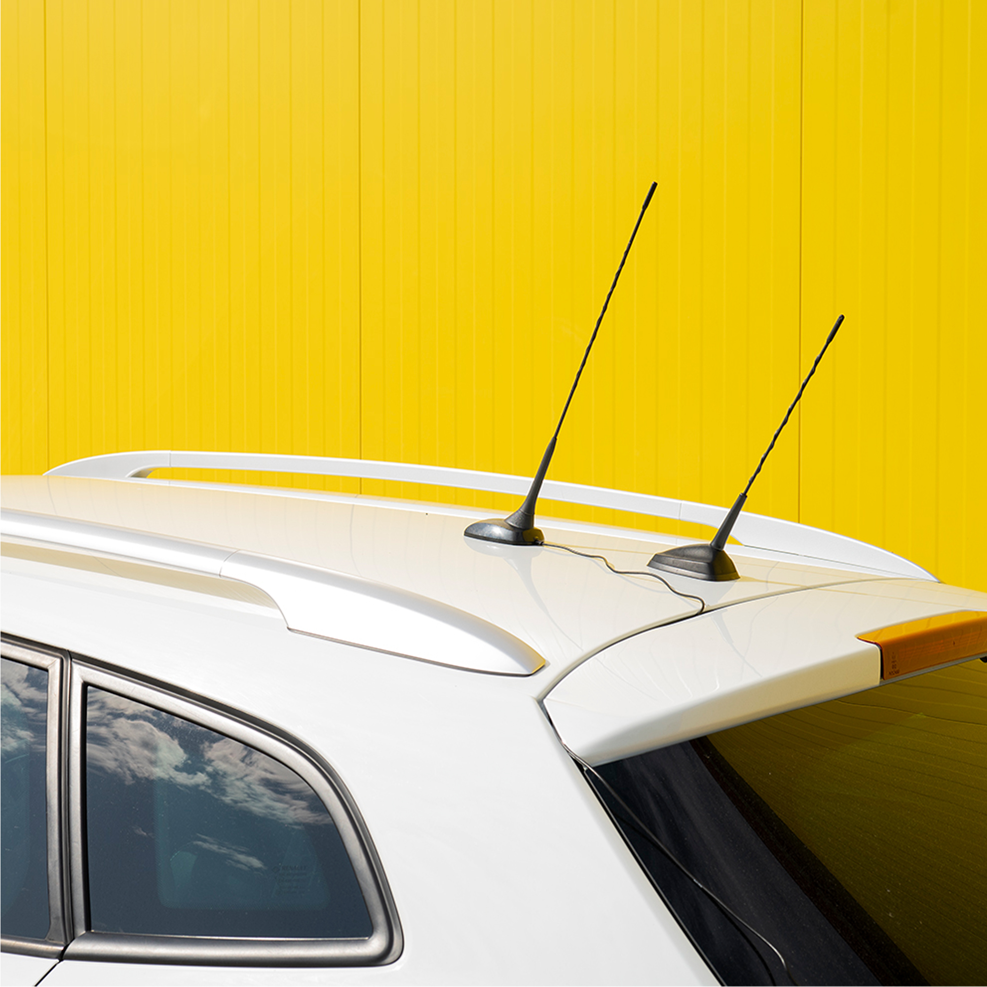 Weißes Autodach mit zwei Antennen vor einer leuchtend gelben Wellblechwand.
