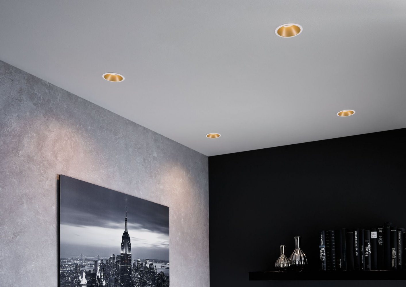 Scatto di interni con luci dorate a incasso nel soffitto, parete grigia e parete nera con arte.