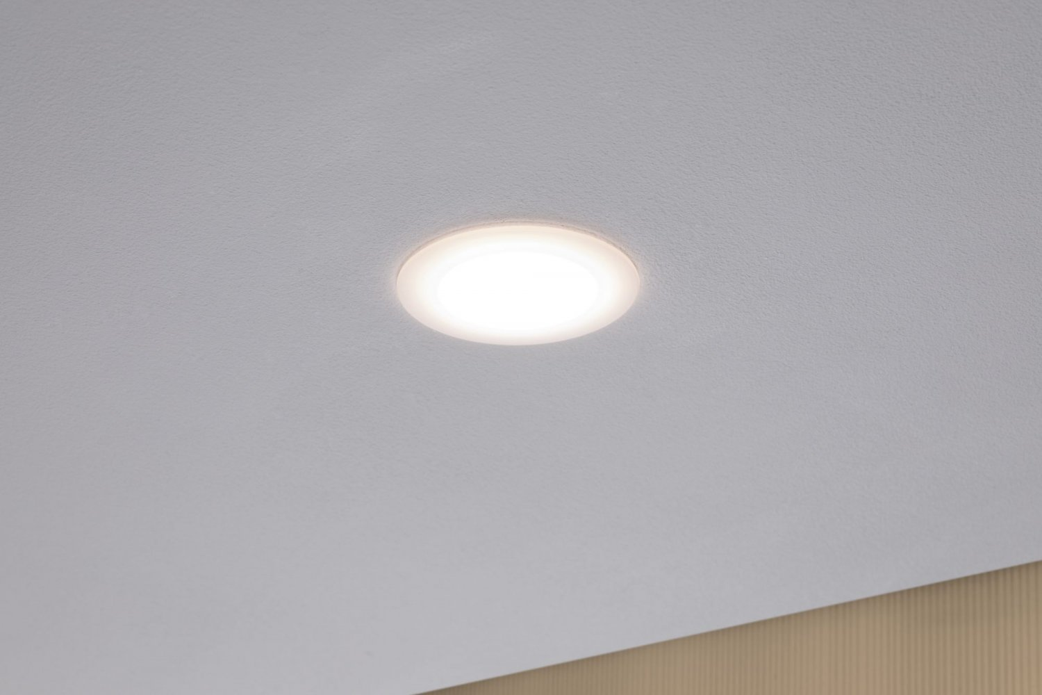 Eine Einbau-Deckenleuchte ist eingeschaltet. Der Hintergrund ist schlicht weiß und eine hellbraune Wand.