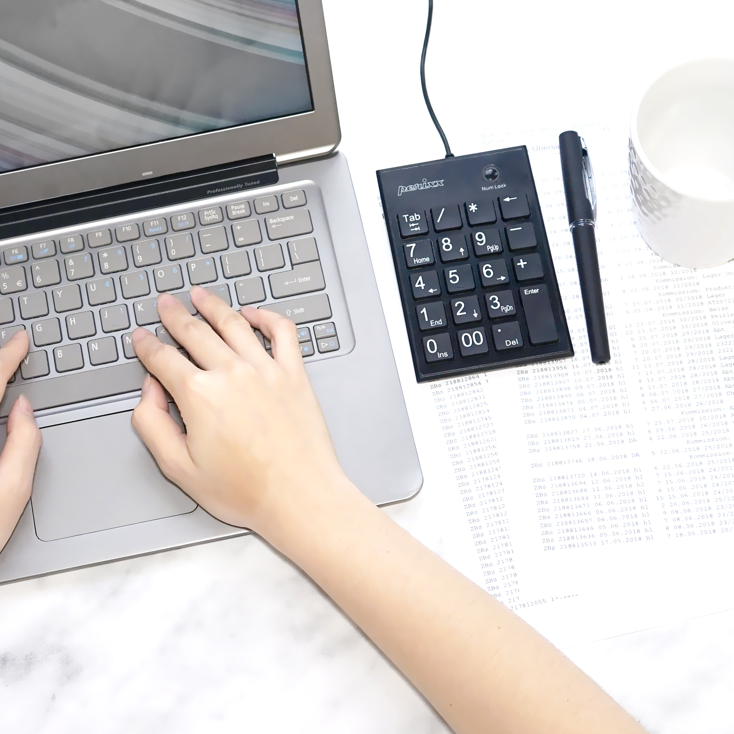 Hände, die auf einem Laptop tippen, neben einem Ziffernblock und Papieren mit Text, einem Stift und einer Tasse.