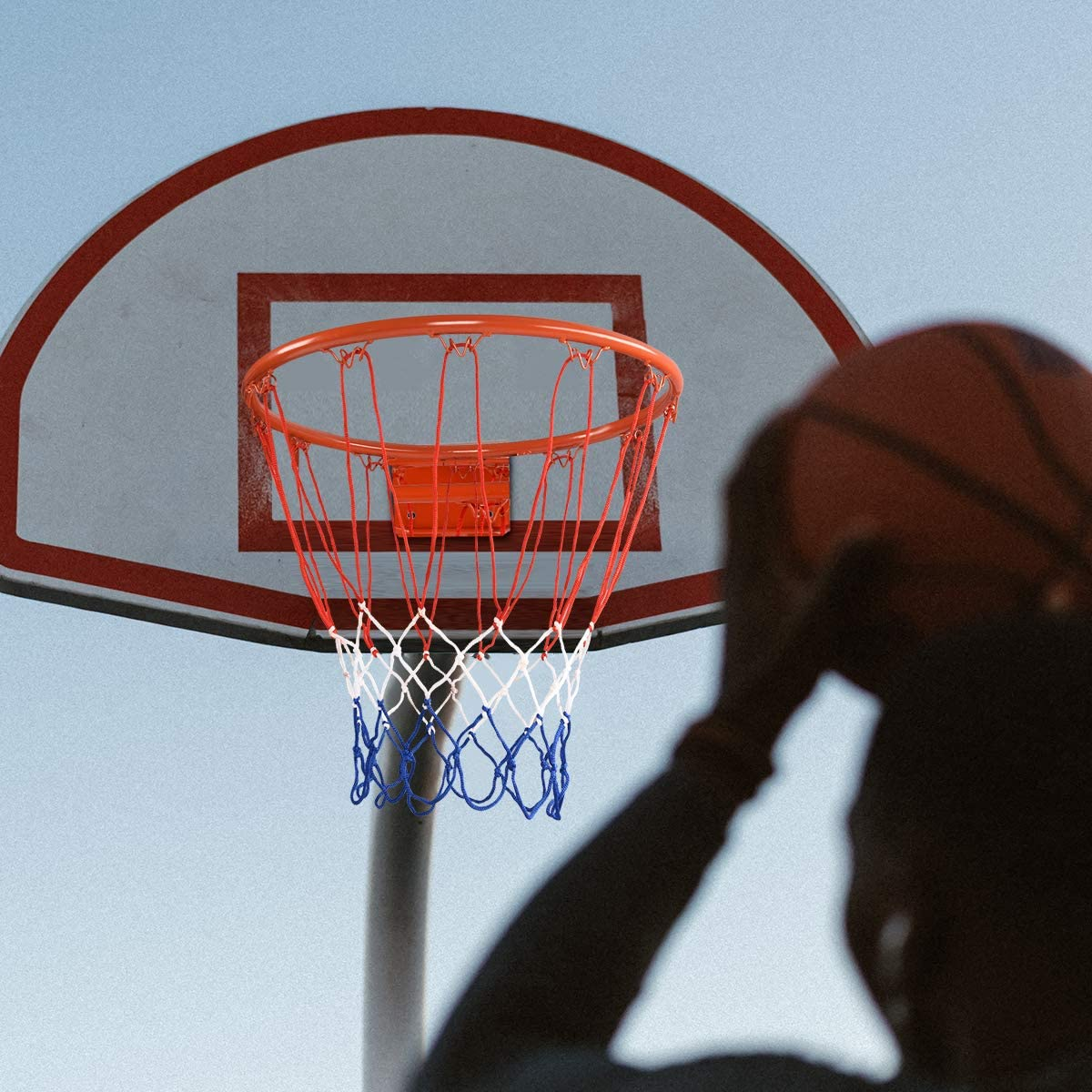 Ein Basketballkorb mit orangefarbenem Rand, rotem, weißem und blauem Netz, und eine Person im Hintergrund als Silhouette.