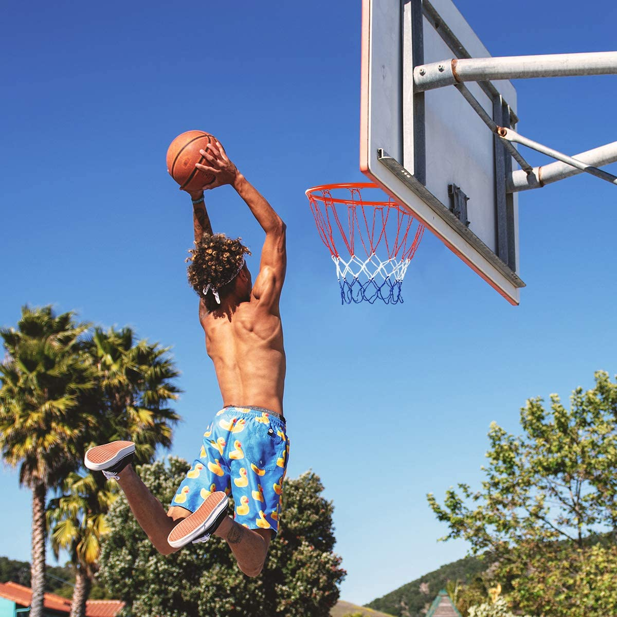 Ein Mann springt, um einen Basketball zu dunken. Er trägt Badehosen. Klarer blauer Himmel und Basketballkorb im Hintergrund.