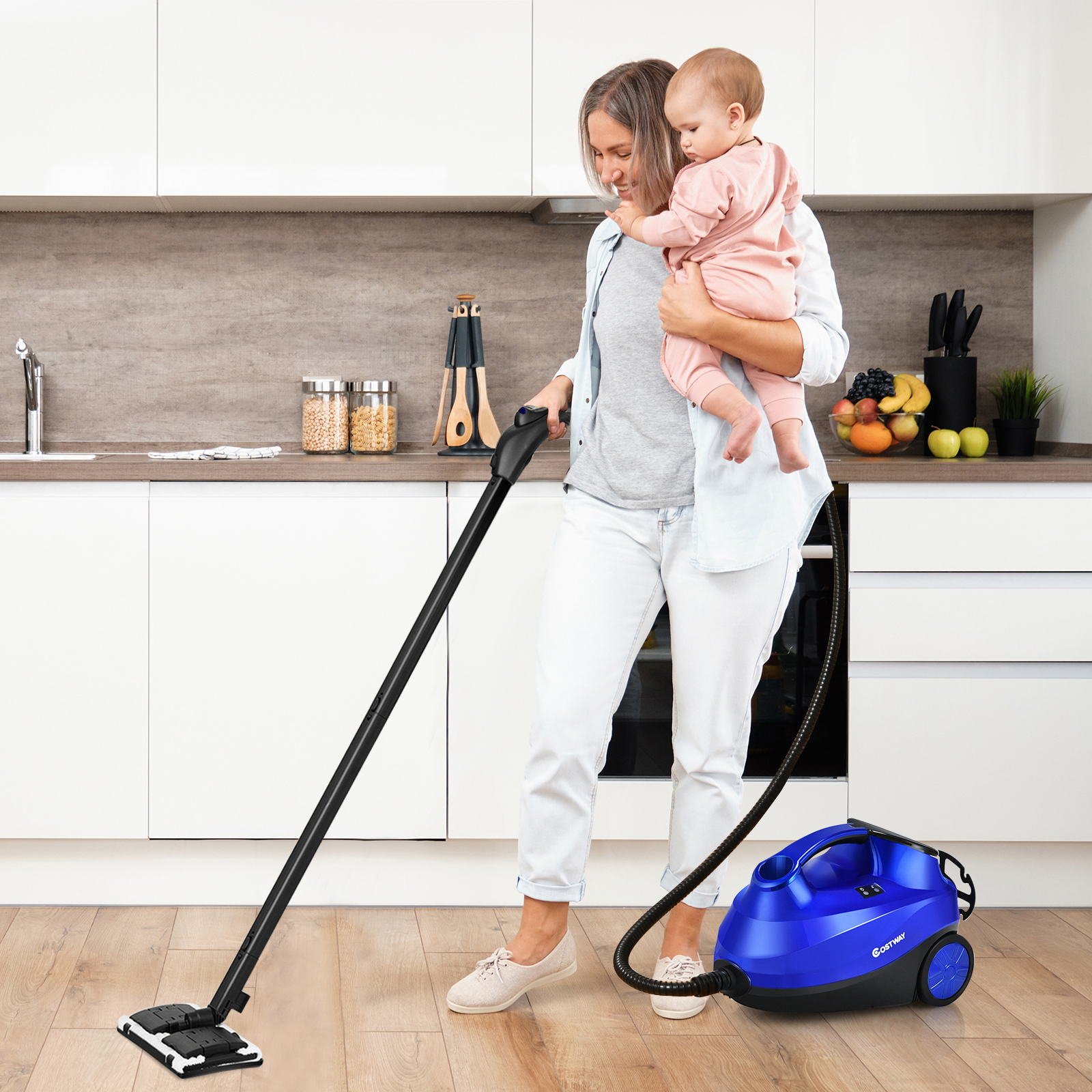 Eine Frau hält ein Baby, während sie den Boden mit einem Dampfreiniger reinigt. Weiße Schränke sind hinter ihnen.