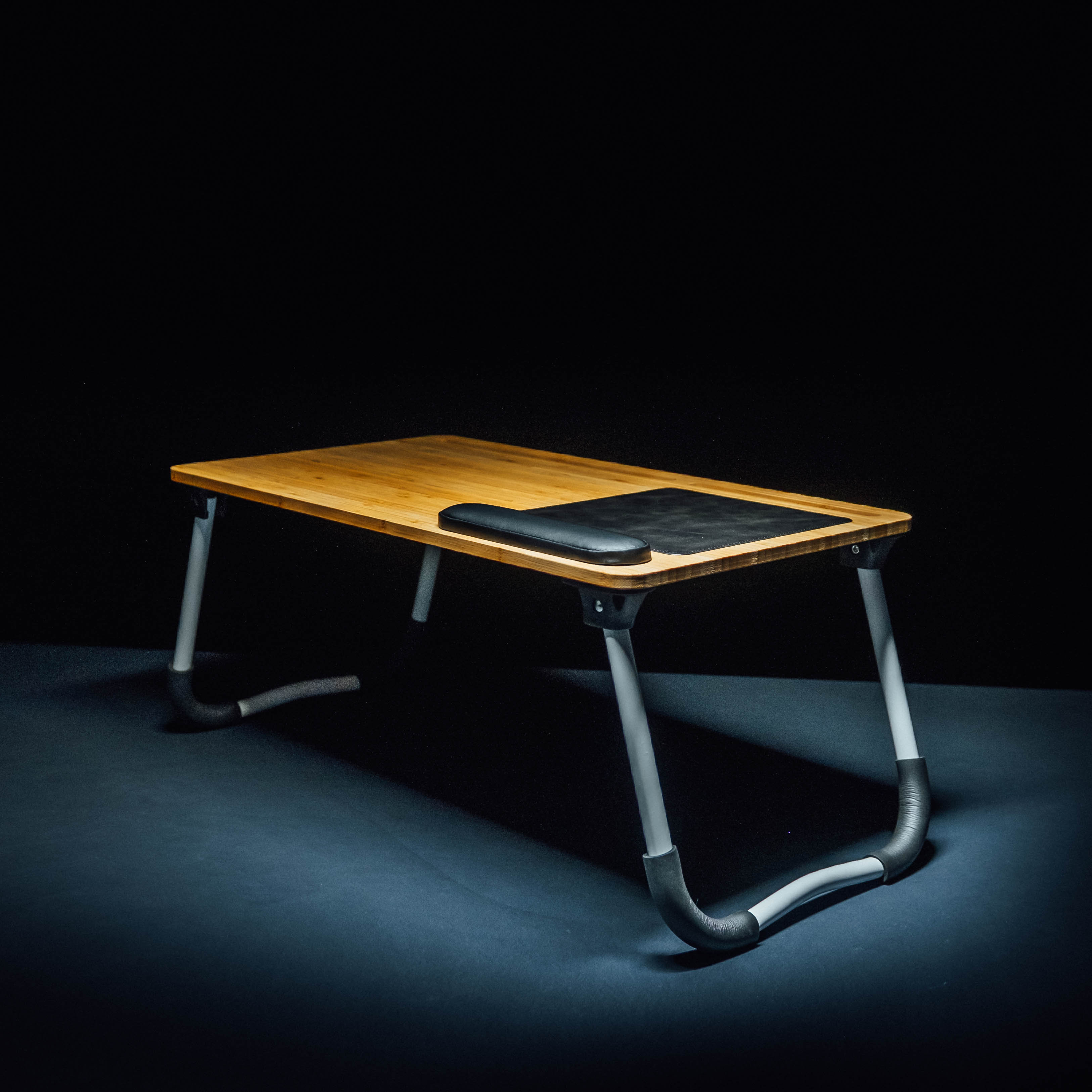 Ein Laptop-Tisch aus Holz mit grauen Metallbeinen. Ein schwarzes Mauspad befindet sich auf der Tischoberfläche. Dunkler Hintergrund.