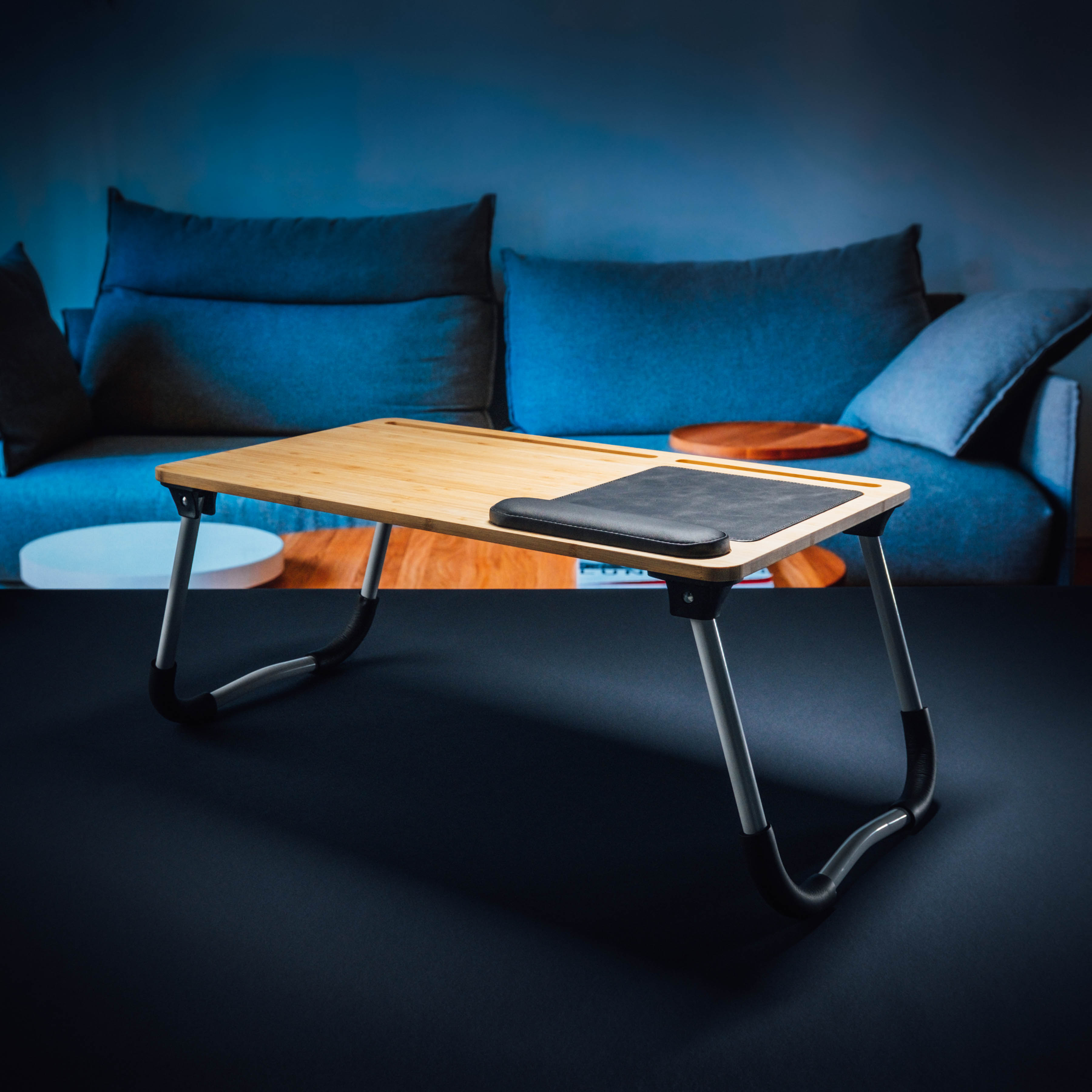 Ein hölzerner Schoßtisch mit einem schwarzen Pad darauf. Er steht vor einem blauen Sofa. Links ist ein weißer Ständer.