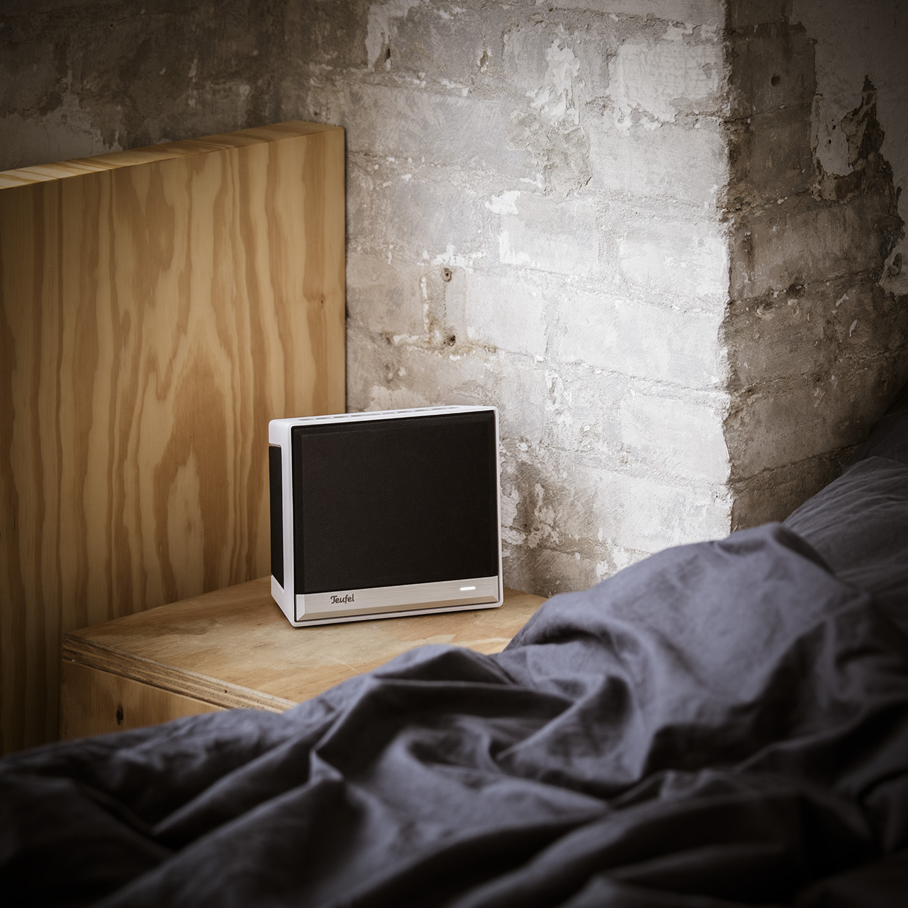 Ein Lautsprecher steht auf einem Nachttisch aus Holz in einem Raum mit einer Backsteinmauer. Das Bett ist rechts.