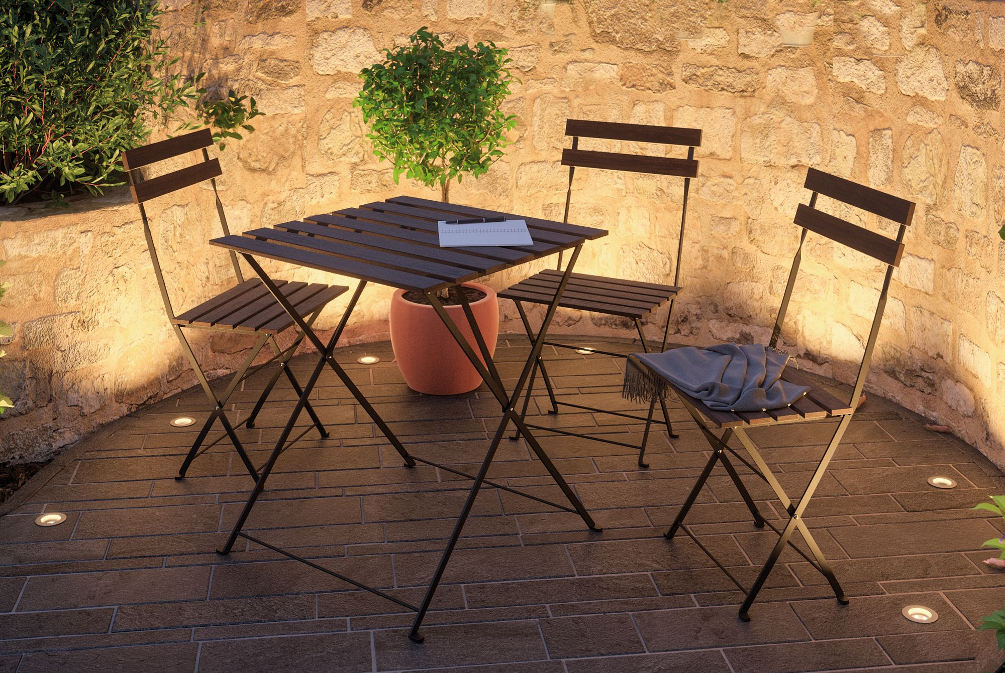 Scena na zewnątrz ze stołem, krzesłami i małym drzewem, oświetlona przez światła gruntowe i kamienny mur.