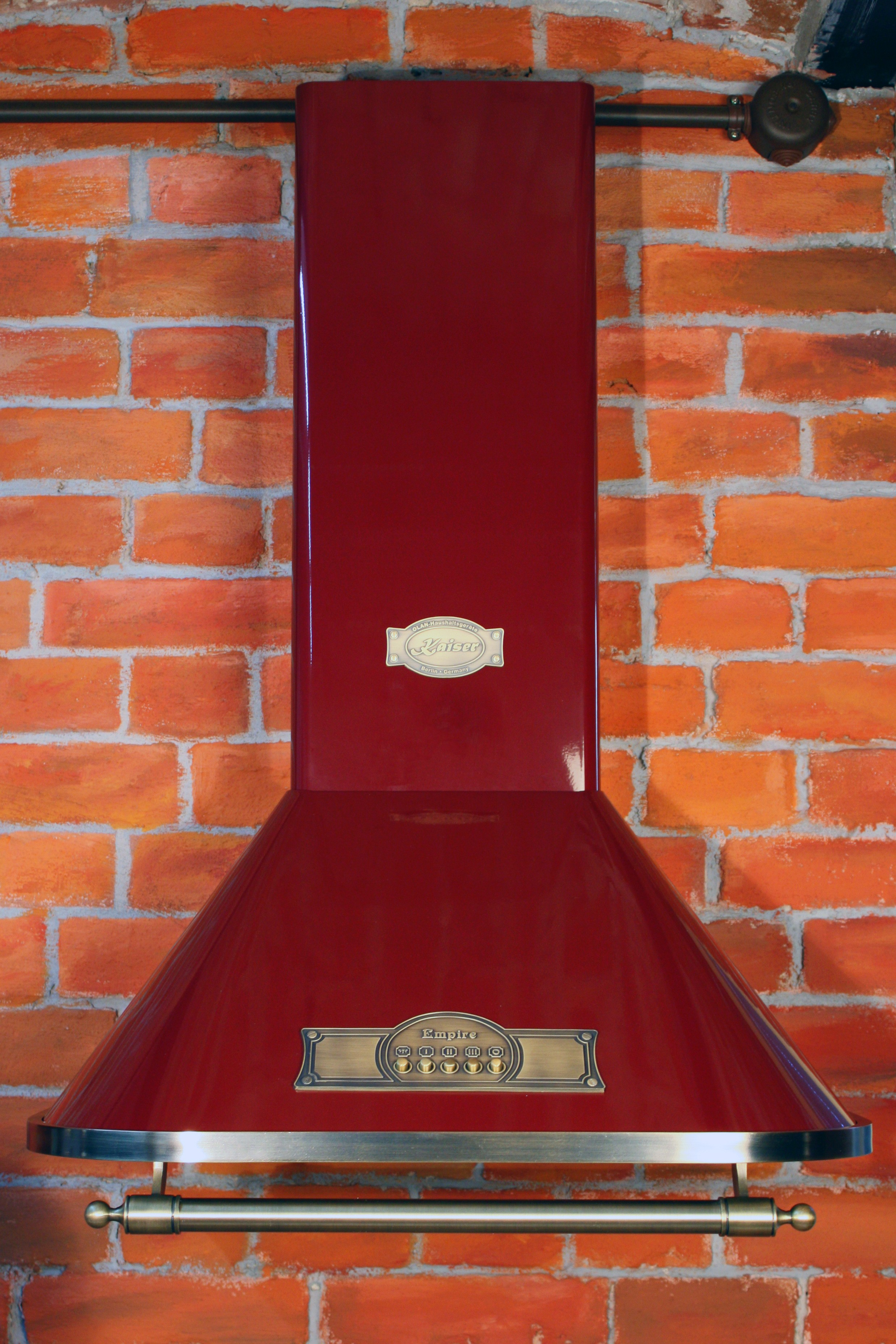 Rote Dunstabzugshaube an einer roten Backsteinmauer, mit goldenen Akzenten und zwei Etiketten.