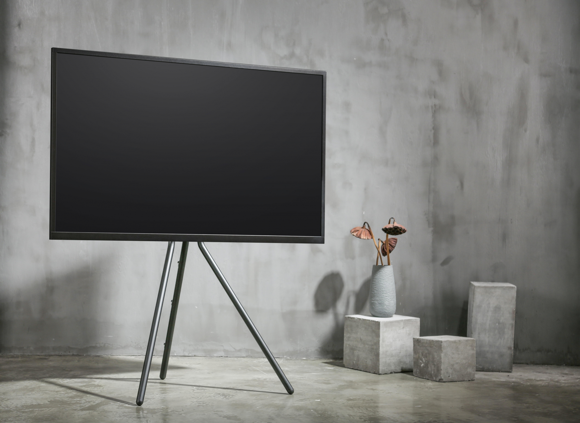 Schwarzer Fernseher auf einem Stativständer. Er steht vor einer grauen Wand mit Betonblöcken.