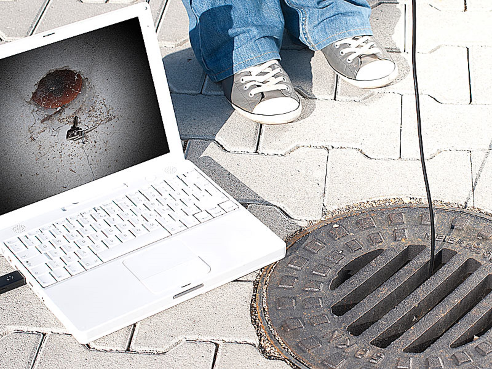 Ein Laptop zeigt ein Kanalinspektionsbild. Eine Person in grauen Schuhen steht in der Nähe auf grauem Pflaster.