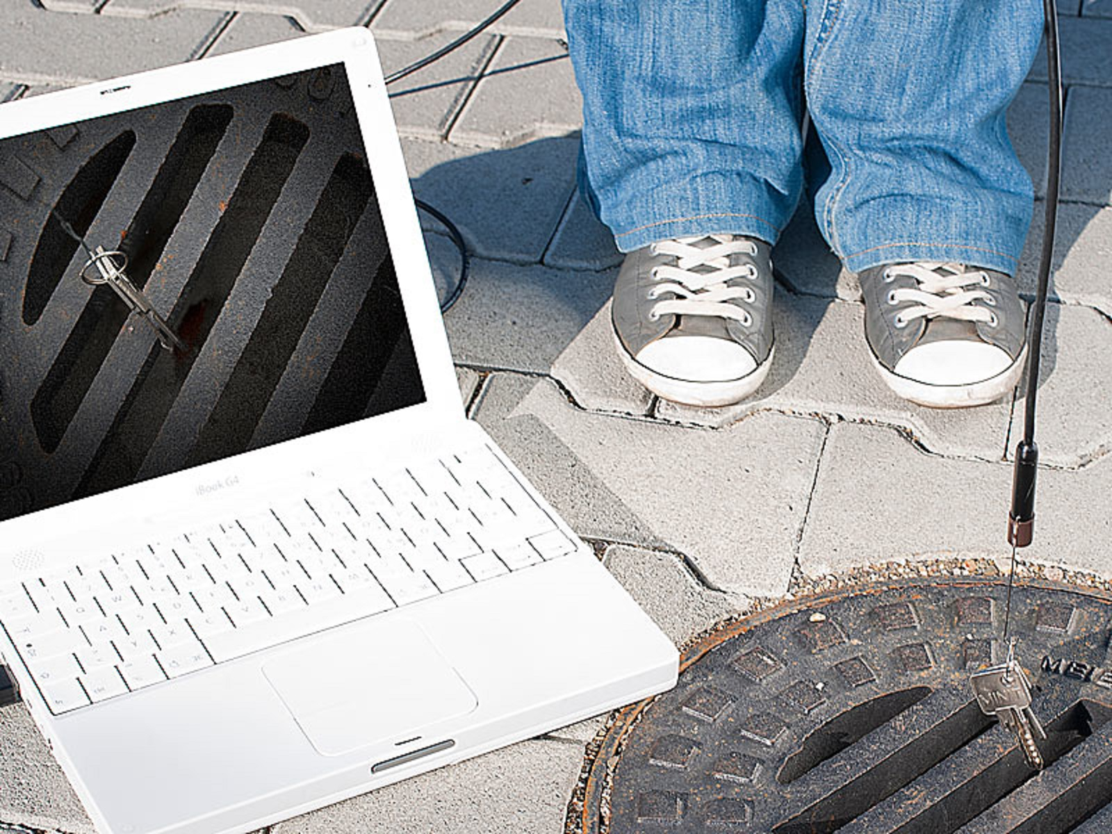 Ein Laptop neben einem Kanaldeckel mit Schlüsseln darin, Beine einer Person im Hintergrund.