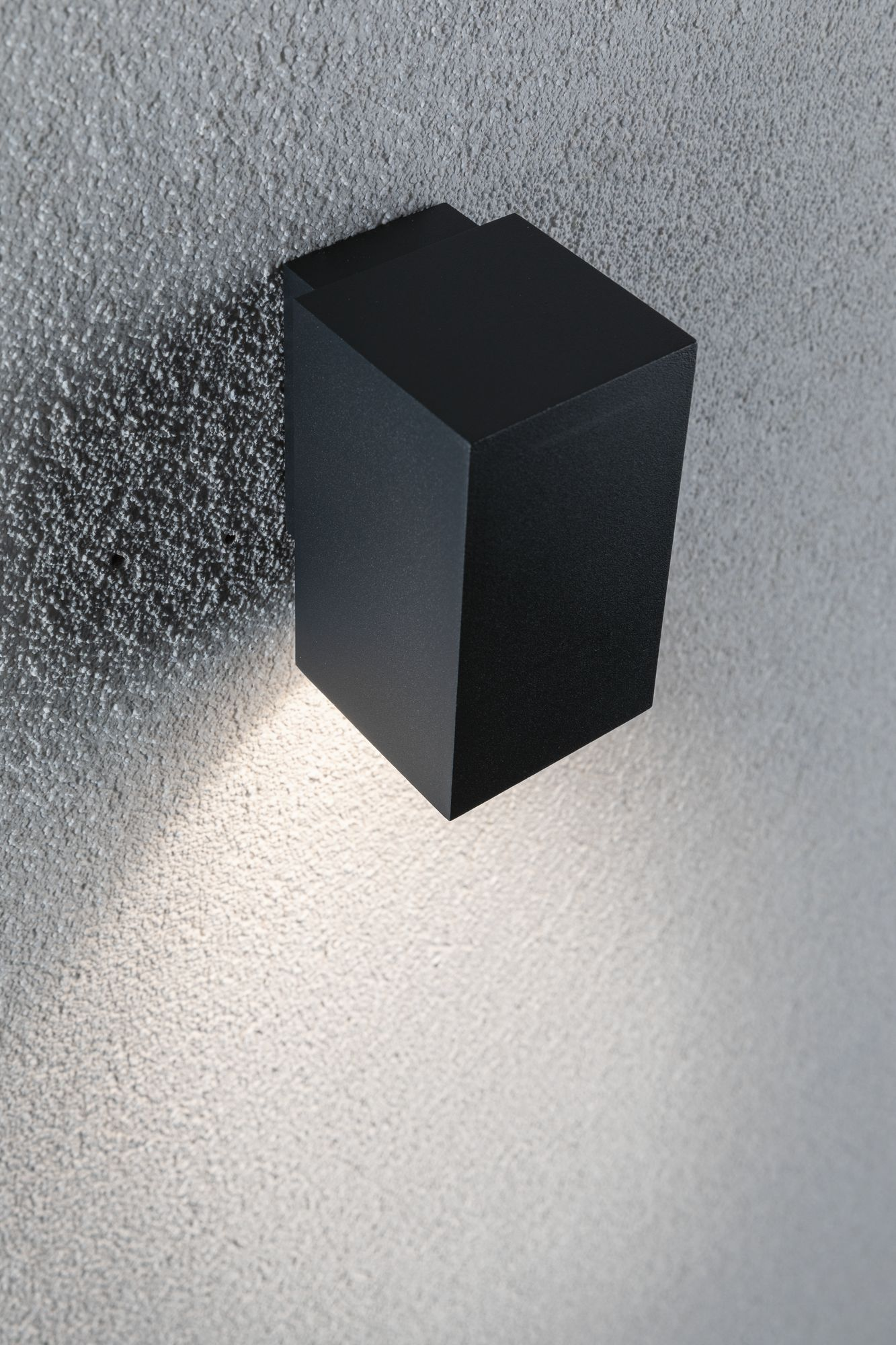 Czarna prostokątna lampa ścienna emitująca światło w dół na ścianę o chropowatej fakturze.
