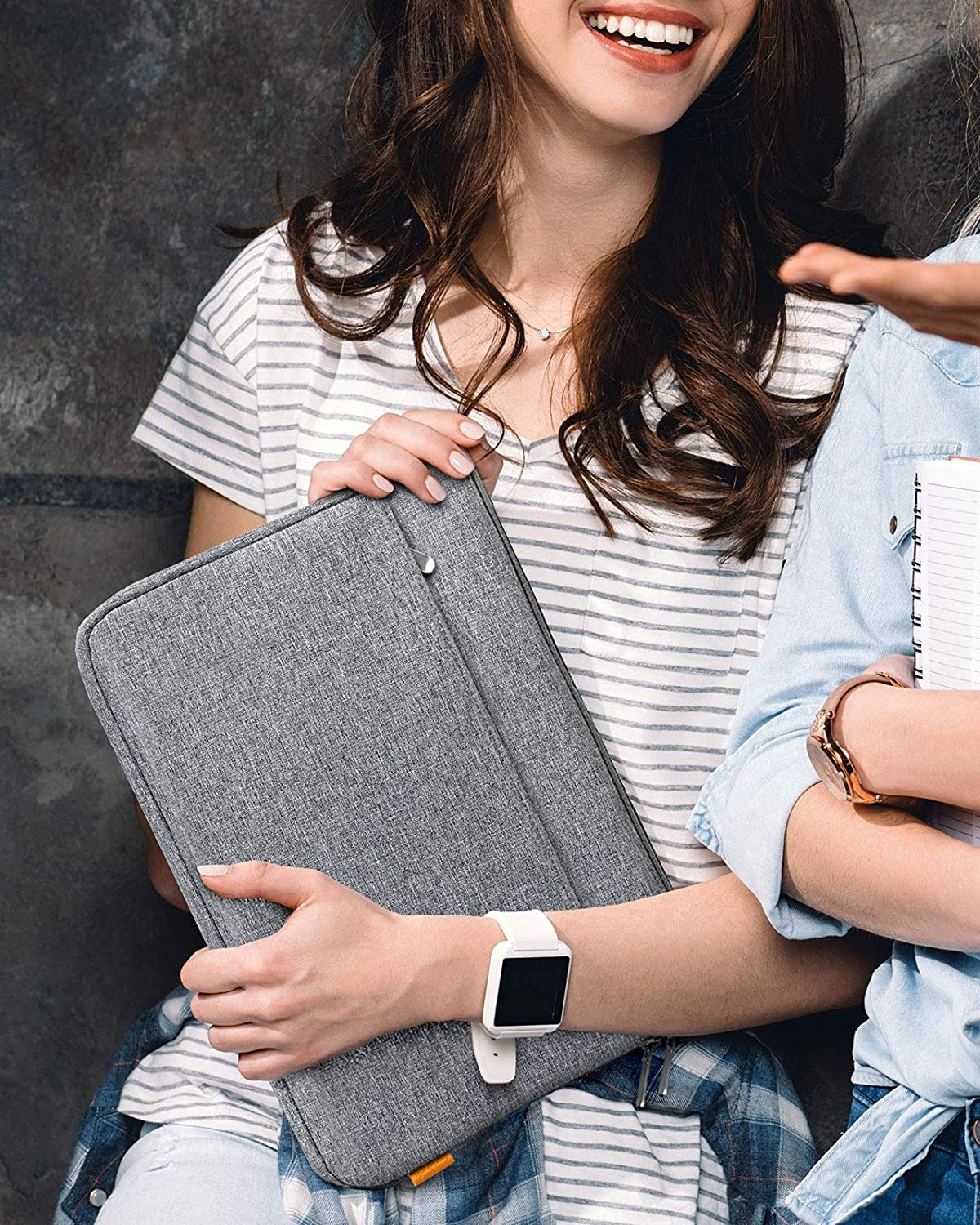 Frau hält graue Laptoptasche, trägt gestreiftes Hemd und weiße Uhr. Eine andere Person ist rechts.