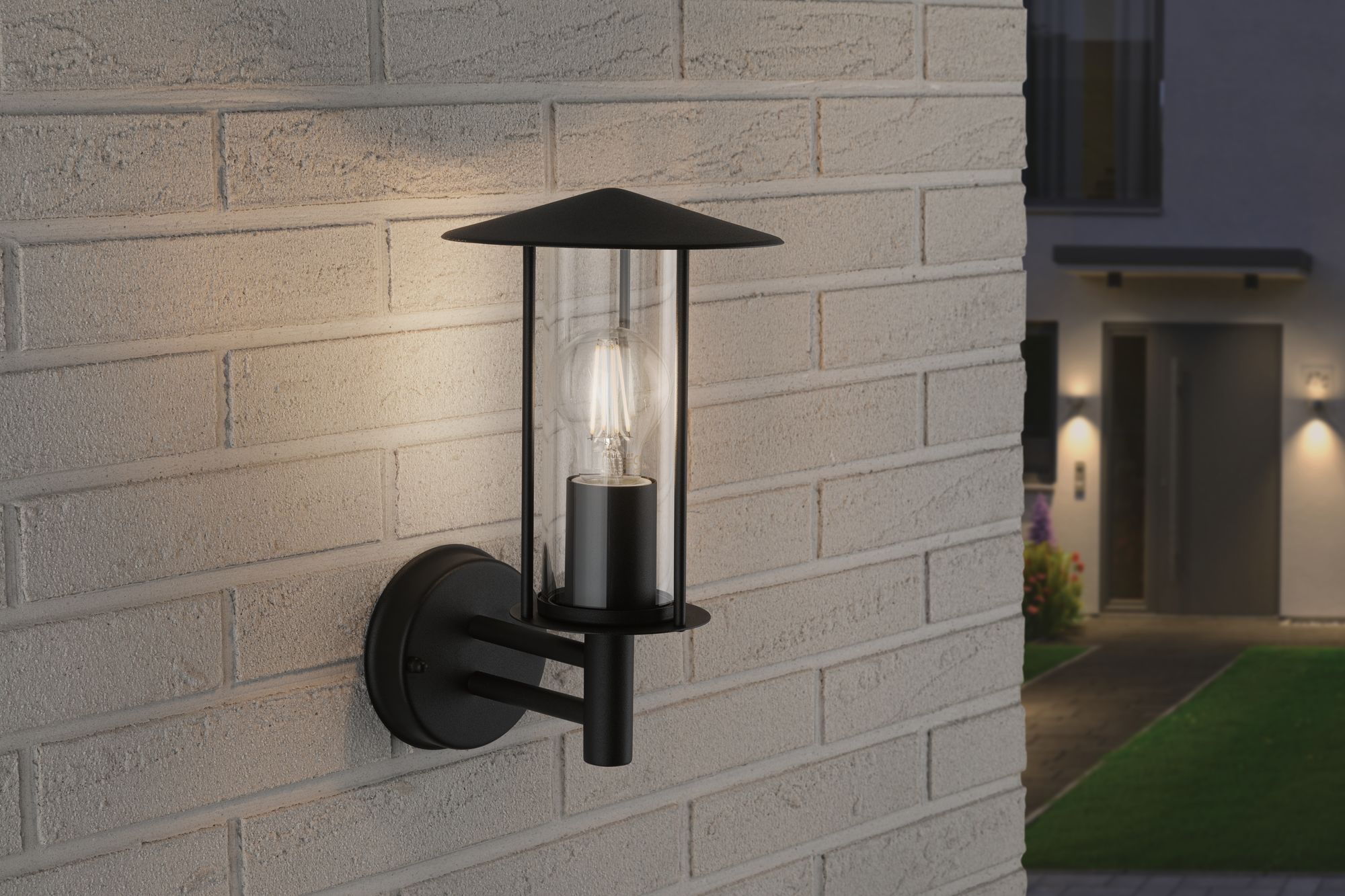 Zewnętrzna lampa ścienna na ceglanej ścianie. Lampa ma czarną ramę i szklaną obudowę.