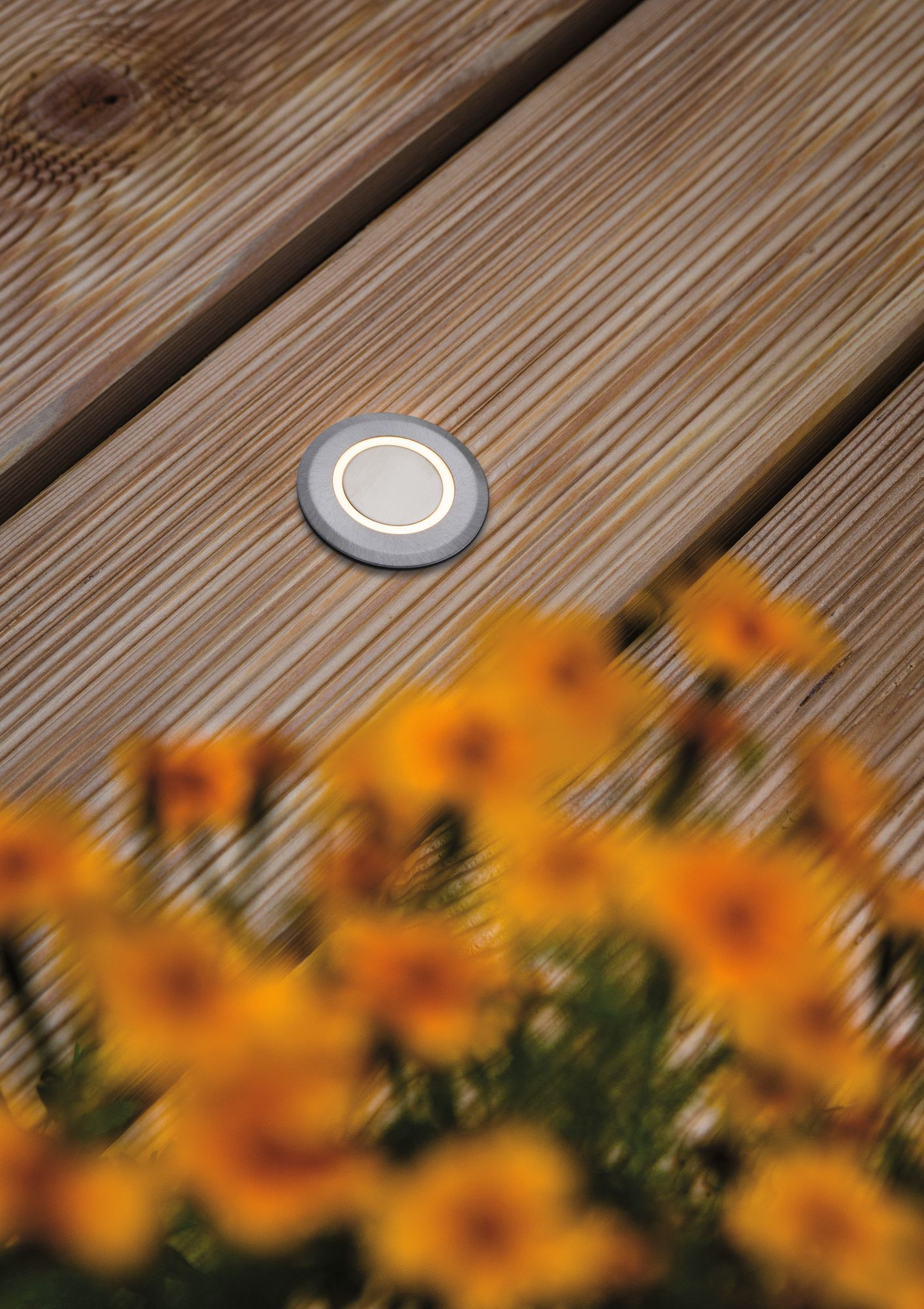 Mała, okrągła, metalowa lampa zewnętrzna umieszczona w drewnianym tarasie, z rozmytymi żółtymi kwiatami.