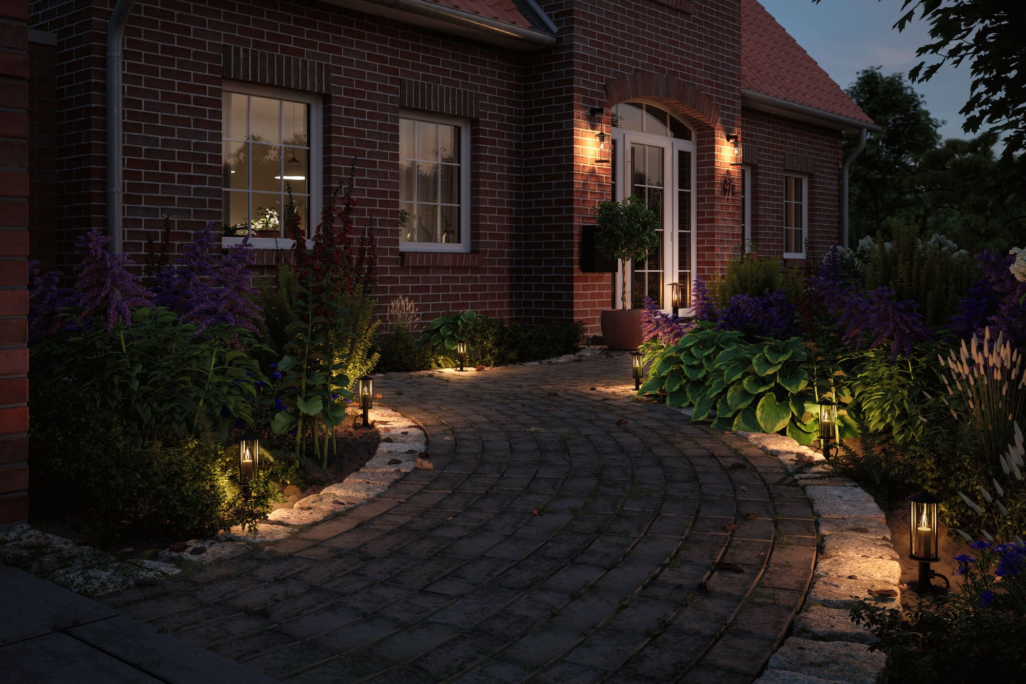 Scatto notturno di una casa in mattoni con vialetto illuminato e luci da giardino, fiori e piante visibili.