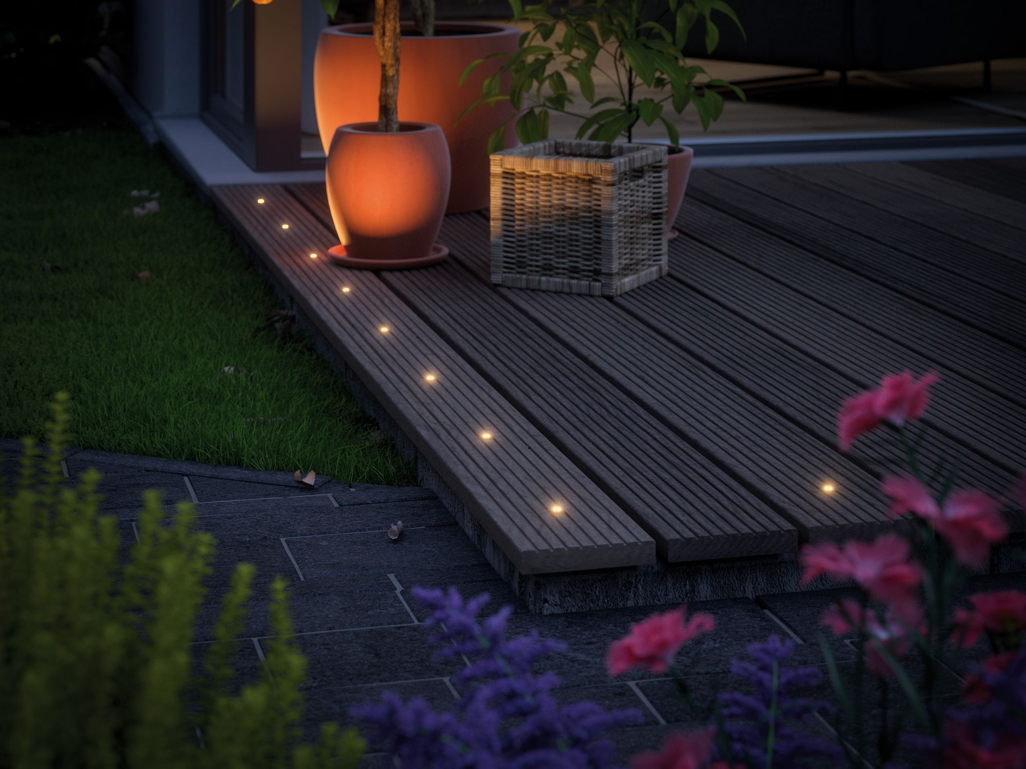Scena all'aperto: terrazza in legno con luci, vasi, piante e fiori. Lo sfondo è un prato e un edificio.