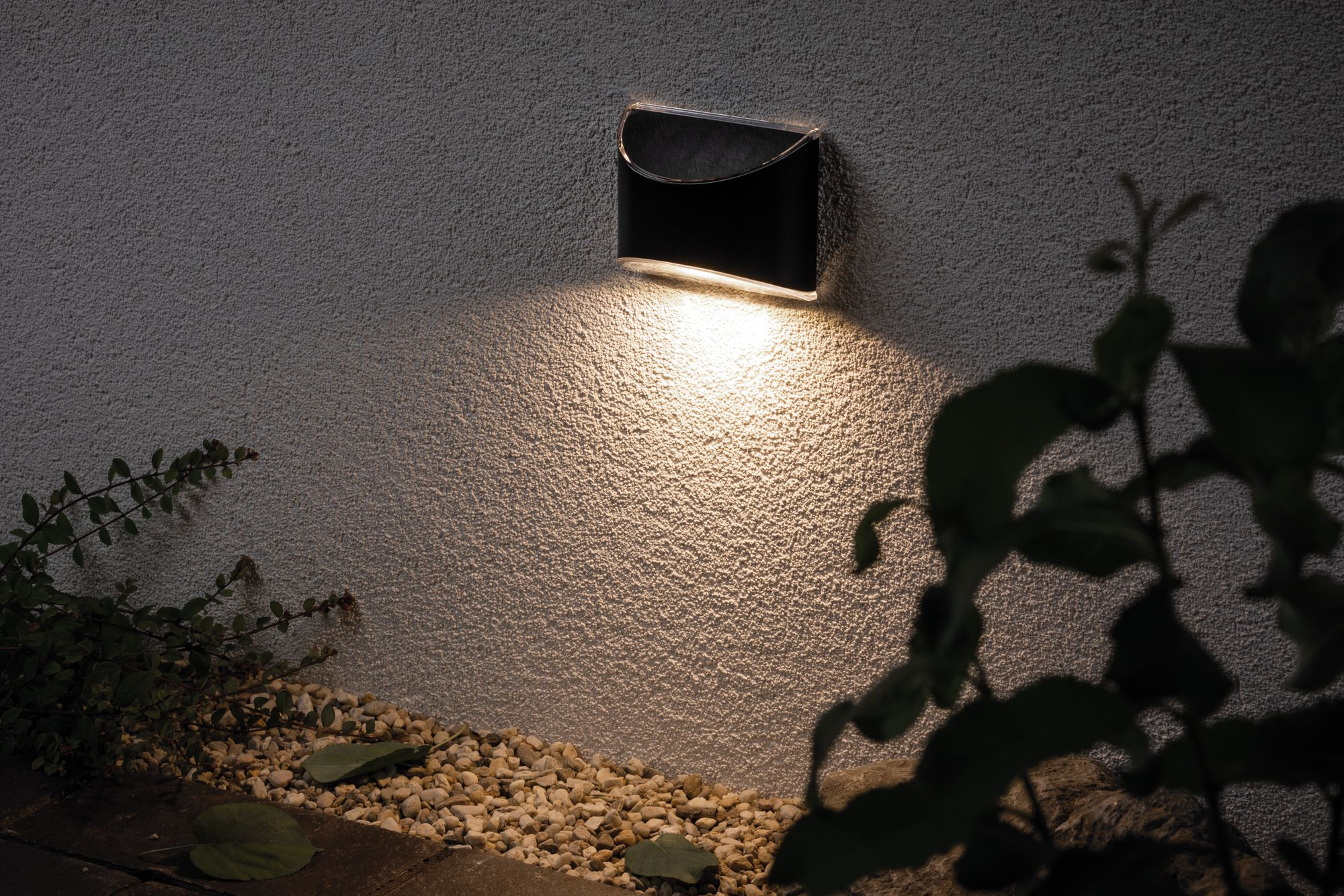Lampada da parete per esterni che illumina un muro. Ciottoli e piante in primo piano.