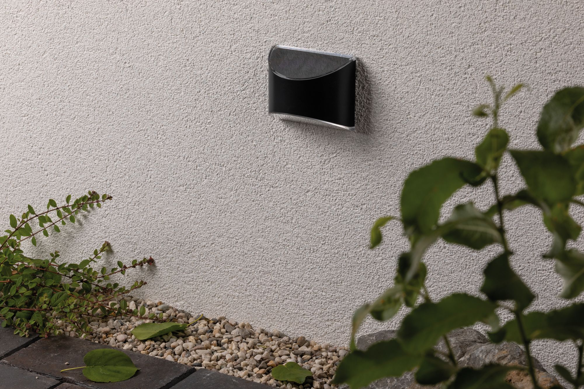 Una lampada da esterno nera su un muro bianco strutturato. Fogliame verde e ghiaia sotto.