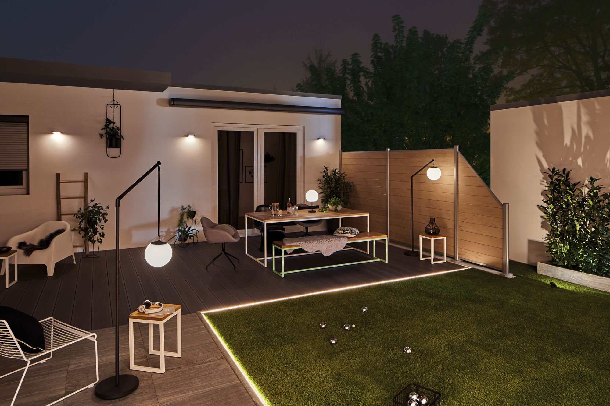 Podwórko nocą ze światłami. Obecne jest drewniane ogrodzenie i meble.