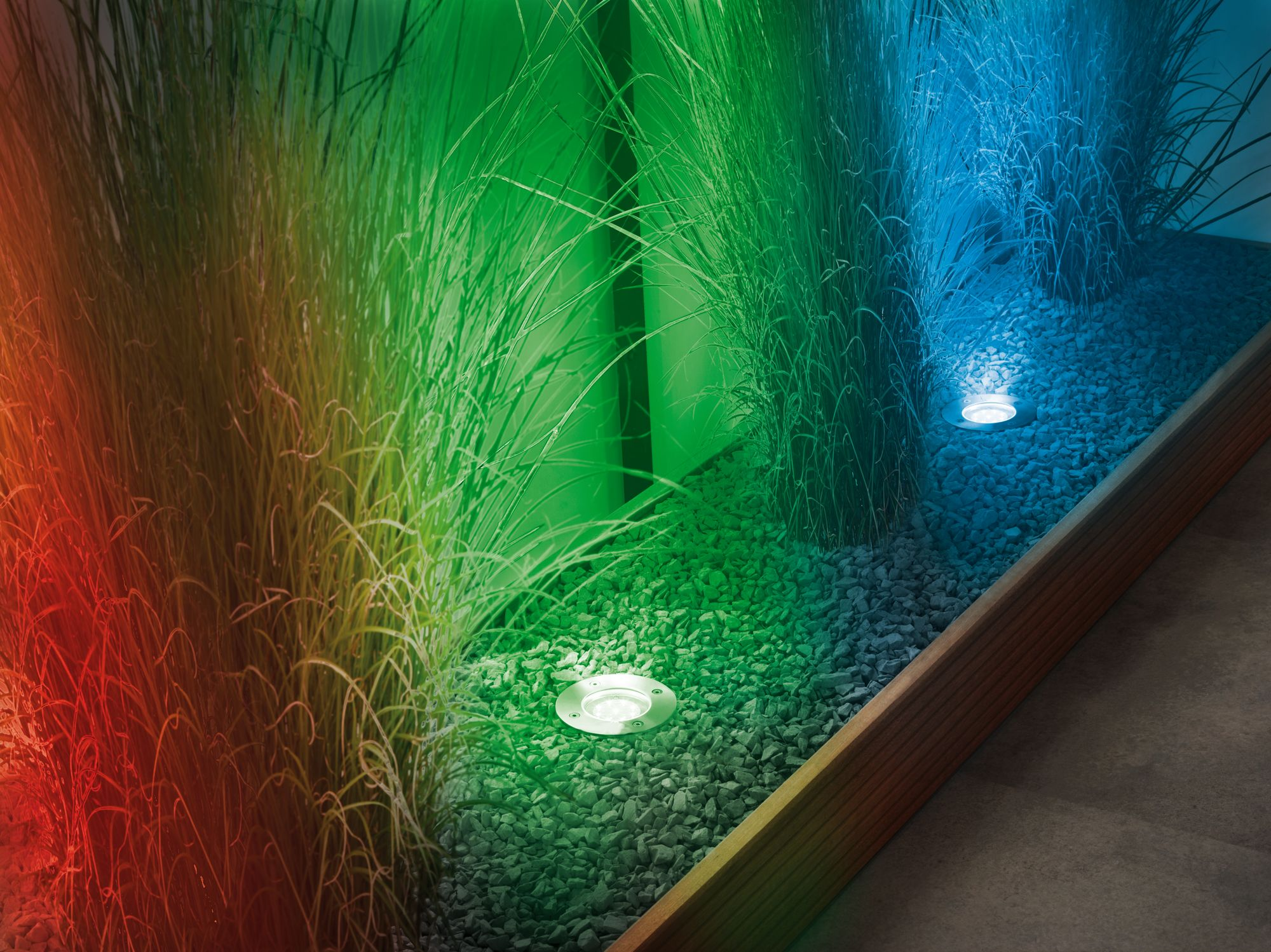 Trzy zewnętrzne reflektory z kolorowymi światłami na trawie i kamykach.