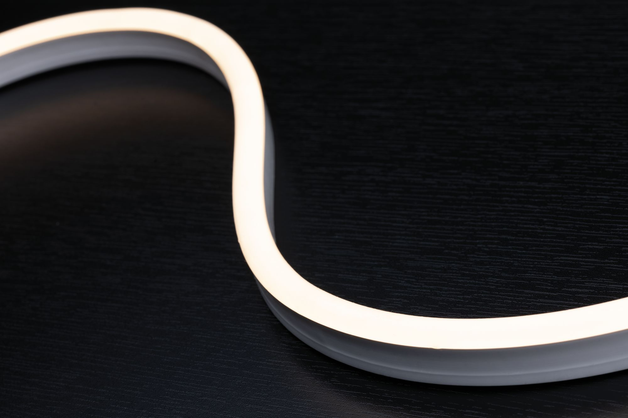 Ein weißer, flexibler LED-Streifen auf einer dunklen Holzoberfläche. Der Streifen ist gebogen und leuchtet.